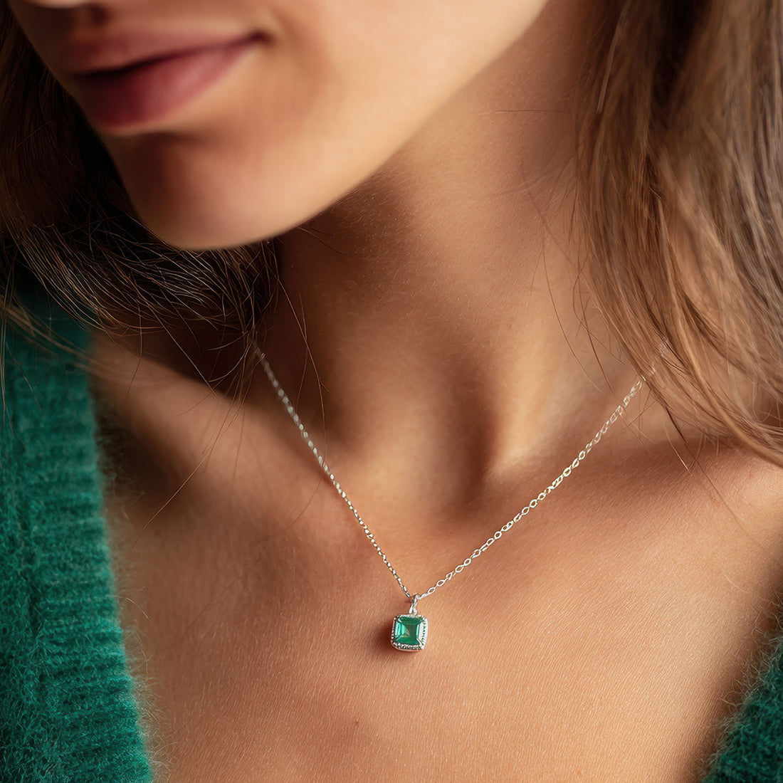 Sieviete ar smalku sudraba krāsas kaklarotu un kvadrātveida kulonu, kurā iestrādāts zaļš akmens.